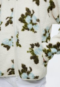 Veste polaire blanche avec des motifs floraux verts et bleus, une fermeture éclair devant et une main placée dans la poche latérale.