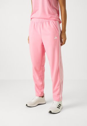 Persoon die roze Adidas trainingsbroek met witte strepen en witte Adidas sneakers draagt op een effen lichte achtergrond.