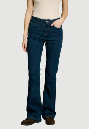 Mujer con vaqueros azul oscuro tipo bootcut, top beige sin mangas, zapatos marrón oscuro, de pie con una mano en el bolsillo frente a un fondo liso.