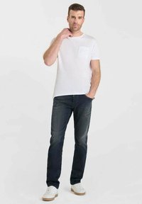 T-shirt blanche à col rond avec une poche poitrine, associée à un jean bleu foncé et des baskets blanches. Design simple, coupe décontractée, sans motifs visibles.