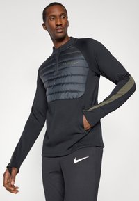 Top sportiv negru cu un piept căptușit albastru, guler cu fermoar și accente verzi măslinii. Asortat cu pantaloni negri care au un logo alb.