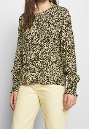 Kobieta ubrana w czarną bluzkę z długim rękawem w drobny żółty kwiatowy wzór oraz kremowe spodnie, stojąca na jednolitym tle.