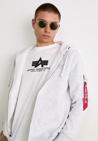 Grå zip-up hættetrøje med frontlommer, lagdelt over en hvid T-shirt med et sort logo. Rød mærkat på ærmet siger "FJERN FØR FLYVNING."