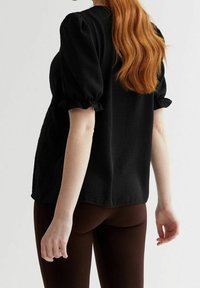 Haut noir à manches bouffantes en tissu texturé, avec un ourlet arrondi, porté avec des leggings marron. Cheveux lisses tombant dans le dos.