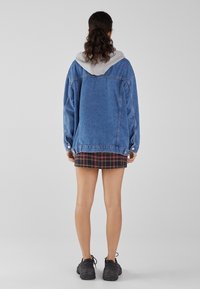 Spijkerjasje met een grijze capuchon, losse pasvorm en knoopdetails. Draagt het over een geruite rok. Zwarte sneakers zichtbaar aan de onderkant.