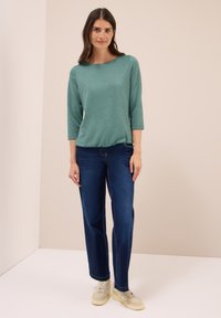 Vrouw staat in een teal 3/4-mouwen top, donkerblauwe rechte jeans en beige sneakers voor een effen beige achtergrond.