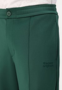 Roheline veniv kangast püksid, millel on nup closure, küljetaskud ja tikitud logo detail. Kitsas lõige struktuurse kujundusega elementidega.