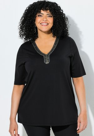 Femme souriante aux cheveux noirs bouclés portant un haut noir à manches courtes avec un décolleté en V orné de perles sur fond blanc.