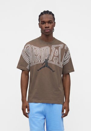 Ung mand med flettet hår iført en brun oversized Jordan t-shirt med store hvide bogstaver og et sort Jumpman-logo, kombineret med lyseblå shorts.