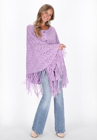 Lila stickad poncho med öppen vävdesign, med franskanter och tre knappar. Parat med ljusblå utsvängda jeans och beiga skor.