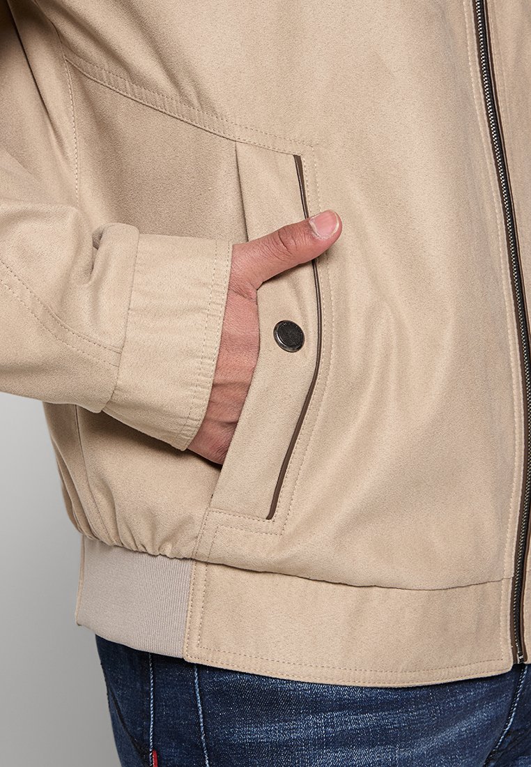 Main placée dans la poche avant d'une veste en daim beige avec bouton-pression et fermeture éclair, associée à un jean en denim bleu foncé.