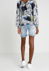 Personne portant un chemisier floral bleu et blanc, un short en denim léger retroussé et des baskets blanches, se tenant de dos sur un fond uni.