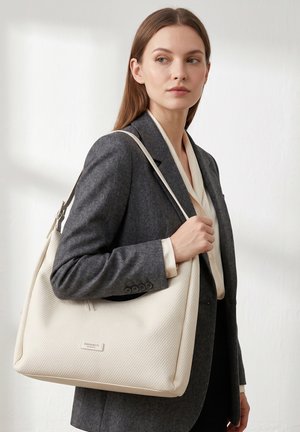 Femme en blazer gris et blouse blanche tenant un grand sac à épaule crème portant l’inscription « DIANA&CO Firenze » sur un fond uni.