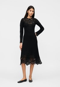 Γυναίκα που φοράει μαύρο midi φόρεμα με μακρύ μανίκι και δαντελωτές λεπτομέρειες στο πάνω μέρος του στήθους, τα μανίκια και το τελείωμα, συνδυασμένο με μαύρα μπαλαρίνα παπούτσια.