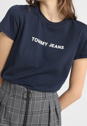 Femme portant un t-shirt bleu marine avec le texte « "TOMMY JEANS" » et une jupe à carreaux grise taille haute avec une fermeture éclair devant et un anneau de tirage.