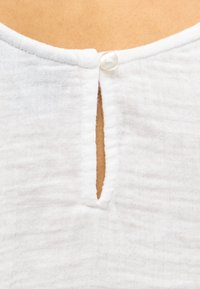 Εγχώριο λευκό υφασμάτινο ένδυμα με λαιμόκοψη, στερεωμένη με ένα μικρό στρογγυλό κουμπί πάνω από κάθετη οπή σε σχήμα κλειδαρότρυπας.