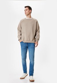 Felpa beige a collo rotondo con polsini e orlo a coste, abbinata a jeans slim-fit azzurri e sneakers bianche con suole marroni.