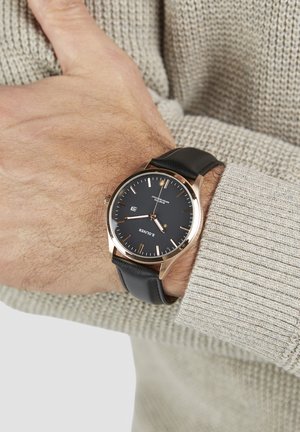 Sort læderrem ur med rundt rosagult etui, mørk urskive, gyldne timemarkører og datovisning. Båret på et håndled med en struktureret sweater.