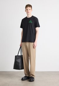 Svart bomull t-skjorte med grønn grafikkdesign på brystet, kombinert med beige bukser og svarte lærsko, holder en totebag.