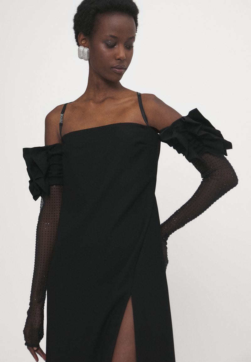 Czarna sukienka bez ramiączek z falbanistymi rękawami, przezroczystymi, kropkowanymi długimi rękawiczkami oraz wysokim rozcięciem, tworząca smukłą, elegancką sylwetkę.