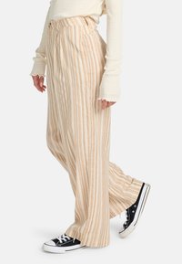 Pantaloni leggeri a gamba larga color beige con strisce arancioni verticali; presentano una vita alta e due tasche laterali, abbinati a scarpe da ginnastica nere.