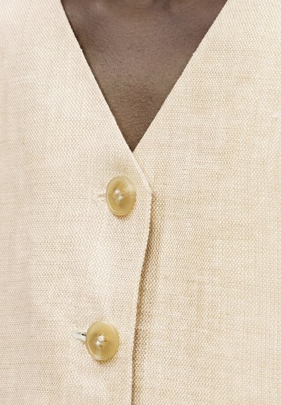 Κοντινό πλάνο μπεζ υφασμάτινου γιλέκου με υφή και δύο διαφανή μπεζ κουμπιά κουμπωμένα πάνω σε σκουρόχρωμο δέρμα.