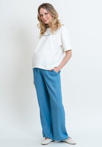 Bílý tričko s krátkým rukávem s nápisem "COOL MAMA", v kombinaci s bledě modrými volnými kalhotami; outfit doplňují bílé pletené boty.