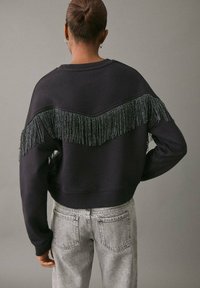 Svart sweatshirt med fransdetalj över den övre ryggen. Funktioner en kort design och ribbade muddar, i kombination med ljusgrå jeans.