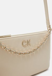 Vaaleanbeige nahkalaukku, jossa on kultasävyinen ketjuolkaiminen, varustettuna vetoketjula closure ja etupuolella kohokuvioitu "CK" -logo. Sujuva pinta, minimalistinen muotoilu.