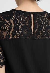 Blusa negra con un yugo de encaje con patrones florales, mangas cortas y un detalle de abertura en la espalda. Tejido suave debajo del encaje.