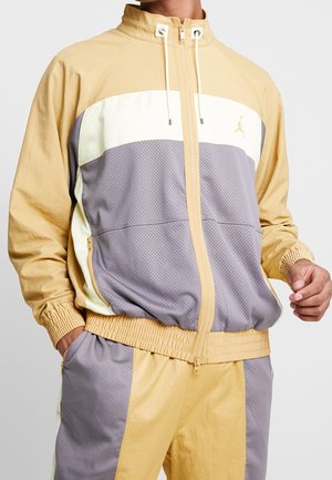 Mężczyzna w beżowo-kremowo-szarej kurtce zapinanej na zamek z blokowym wzorem kolorów oraz pasujących spodniach dresowych z elastycznymi mankietami i kieszeniami.