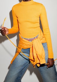 Pull orange côtelé à col montant avec manches longues, associé à un jean en denim bleu clair et une chemise orange boutonnée nouée à la taille.