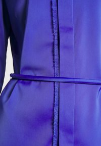 Fialové šaty s hladkým, lesklým materiálem. Obsahují elegantní pásek a roztrhané detaily podél středového švu pro přidanou texturu.