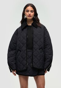 Giacca oversize nera trapuntata con colletto in velluto a coste, chiusura con zip e tasche laterali. Presenta un discreto dettaglio del logo e un tessuto testurizzato.