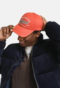 Cappello rosso con testo ricamato "NEW HAVEN Gant" e "CONNECTICUT", indossato con una giacca corduroy blu navy e un interno zip bruno.