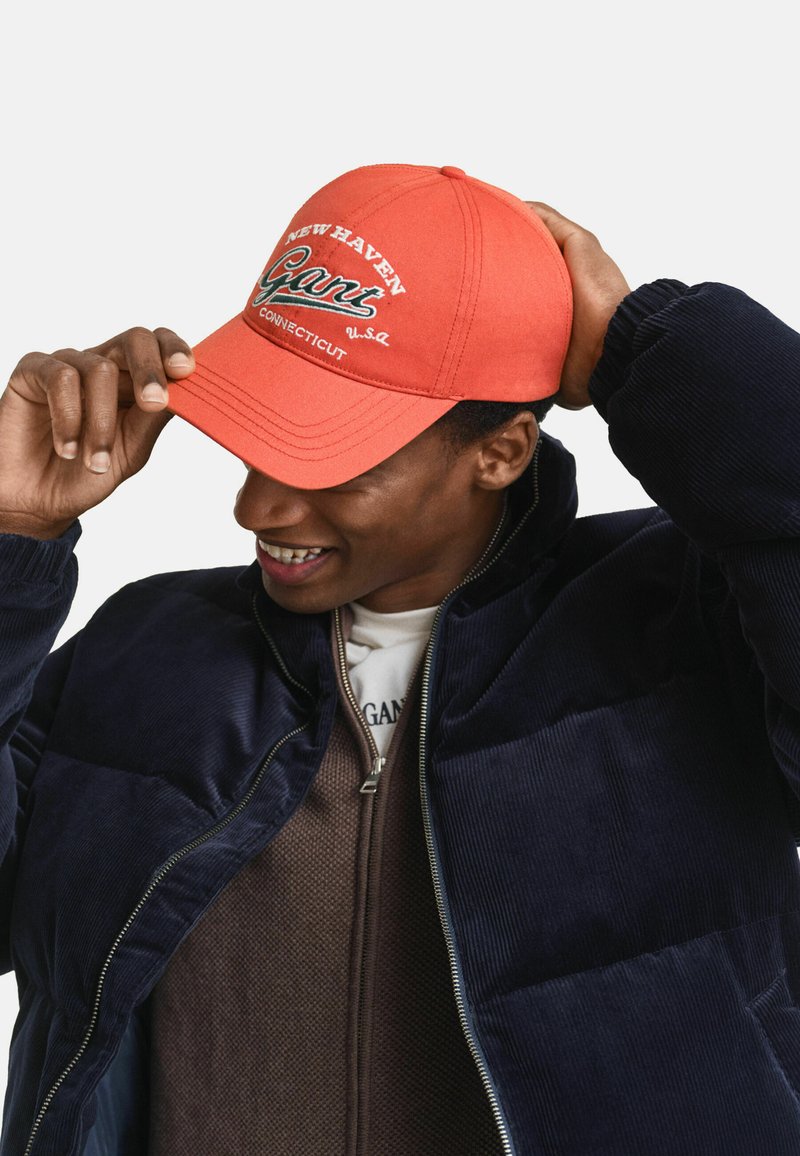 Cappello rosso con testo ricamato "NEW HAVEN Gant" e "CONNECTICUT", indossato con una giacca corduroy blu navy e un interno zip bruno.