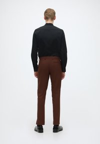 Musta pitkähihaisa paita ja ruskeat muotopuvut nähtynä takaa. Paidassa on suora kaulus; housut ovat slim fit -malliset. Mustat kengät.