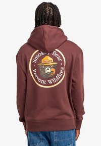 Felpa bordeaux con cappuccio, caratterizzata da una grande grafica circolare di Smokey Bear sul retro. Il materiale sembra morbido, con polsini e orlo a coste.