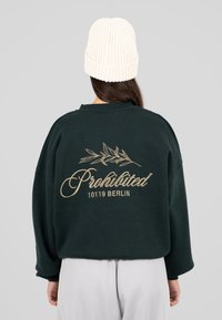 Persona che indossa una felpa verde scuro con il testo beige "Proibito 10119 Berlino" e un disegno di foglia sulla schiena, e un cappellino bianco in maglia.