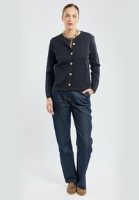 Femme portant un cardigan noir à boutons, un jean bleu foncé, des chaussures à imprimé léopard et un collier en chaîne dorée épaisse, se tenant devant un fond uni.