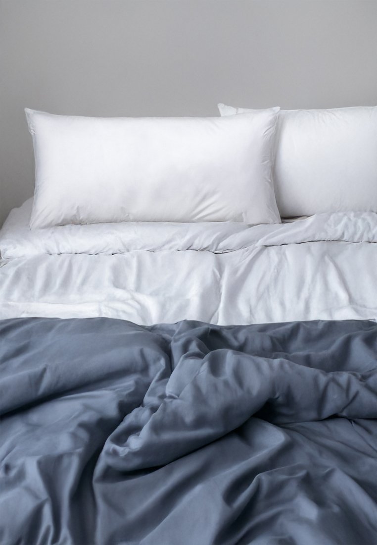 Bettwäsche mit einem weißen Bettlaken und zwei weißen Kissen, bedeckt mit einer zerknitterten dunkelgrauen Bettdecke. Die weiche Stofftextur ist sichtbar.