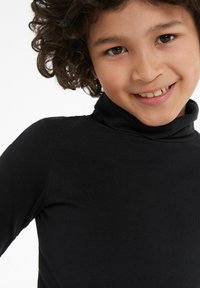 Schwarzer Rollkragenpullover mit einer glatten Textur, tailliertem Schnitt und leichtem Stretch. Das Modell hat lockige Haare und lächelt.