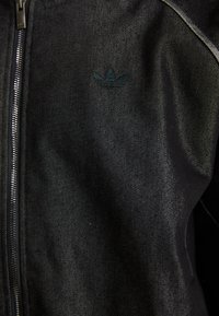 Chaqueta negra con cremallera, hecha de una tela texturizada. Presenta un logotipo oscuro y sutil, así como costuras de contraste a lo largo de las costuras.