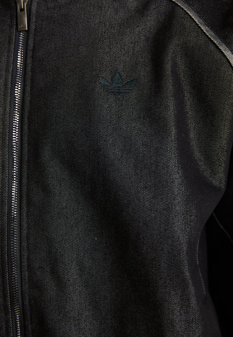 Schwarze Zip-Jacke aus strukturiertem Stoff. Verfügt über ein dezentes, dunkles Logo und Kontrastnähte entlang der Nähte.
