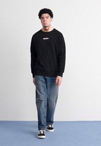 Sudadera negra con logo, jeans de mezclilla azul y zapatillas negras. El modelo se encuentra de pie contra un fondo neutro, mostrando un atuendo casual.