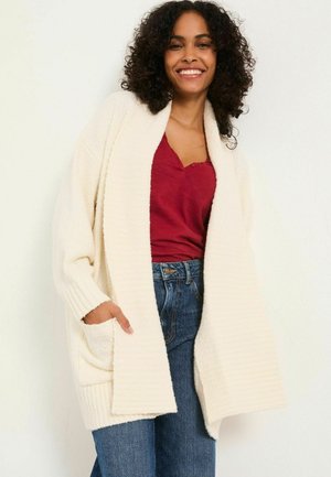 Cremfarbener Cardigan mit gerippter Textur, offener Front und Taschen, getragen über einem burgunderfarbenen Oberteil und kombiniert mit hochtaillierten blauen Jeans.