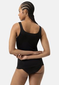 Nainen, jolla on maissikuvioiset letit, pukeutuneena mustaan topiin ja mustiin alushousuihin, seisoo kädet ristissä selän takana, kasvot vasemmalle.