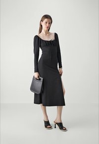 Black long-sleeve dress with a square neckline, tied front, side slit, and textured fabric. Paired with a black handbag and open-toed heels.