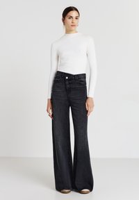 Junge Frau in einem weißen, gerippten Langarmoberteil, kombiniert mit hochgeschnittenen, weit geschnittenen schwarzen Jeans und braunen Schuhen, stehend vor einem schlichten Hintergrund.