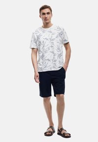 Valkoinen lyhythihainen t-paita, jossa on sininen kukkakuosi, yhdistettynä tummansinsisiin shortseihin ja ruskeisiin sandaaleihin. Yksinkertainen muotoilu, pehmeä kangas ja rento istuvuus.